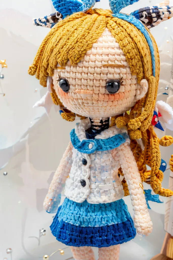 The image shows a handmade doll with long blond hair and blue eyes. She is wearing a white and blue sailor-style outfit with a blue skirt and a white hat. The doll is standing in front of a white background with a star. She is made of yarn and has a wire skeleton.