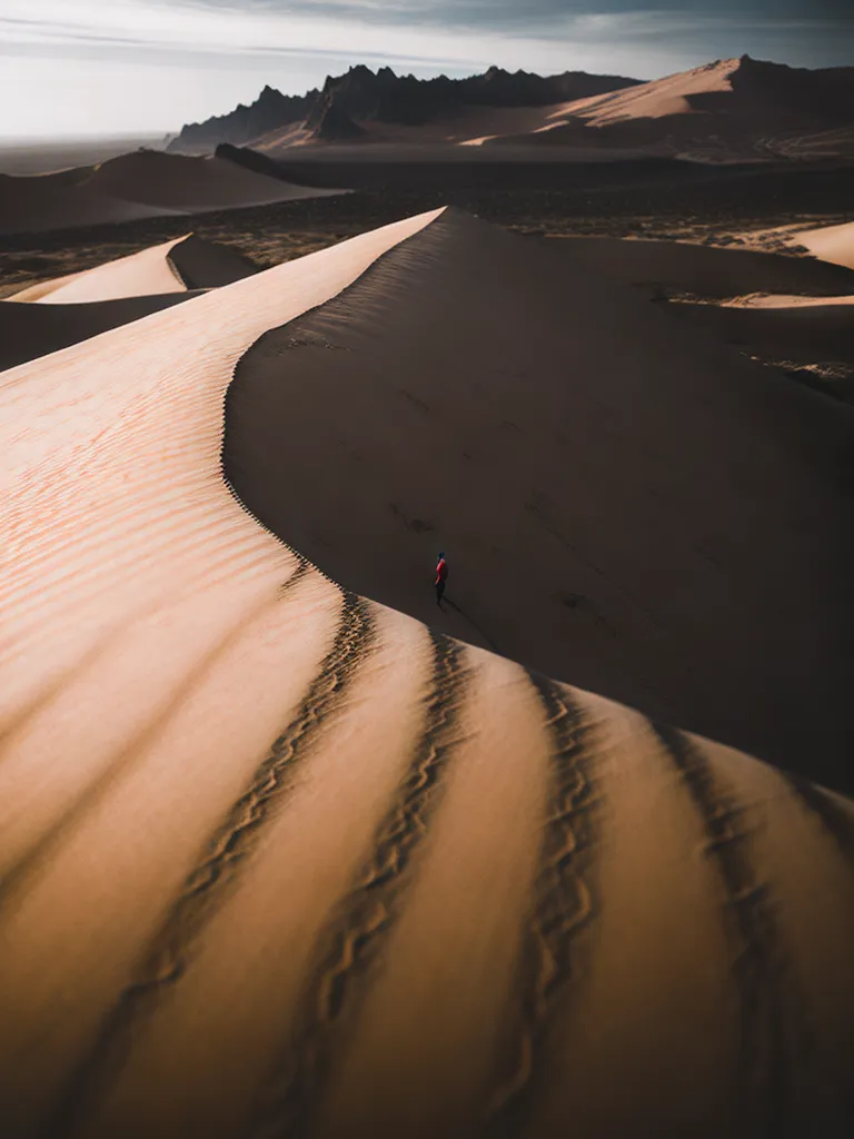 The image shows a vast sand dune in the middle of a desert. The dune is a light golden color and is rippled with footprints. There is a person in a red jacket walking down the dune. They are the only person in the image. The sky is a light blue color and there are some clouds in the distance.