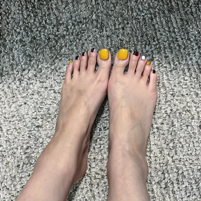 The image shows a person's bare feet with a close-up of their toes. The person's toes are painted with different bright colors. The big toes are painted black with white tips. The second toes are painted white with black tips. The third toes are painted yellow. The fourth toes are painted black with white tips. The fifth toes are painted white with black tips. The person's feet are resting on a gray carpet.