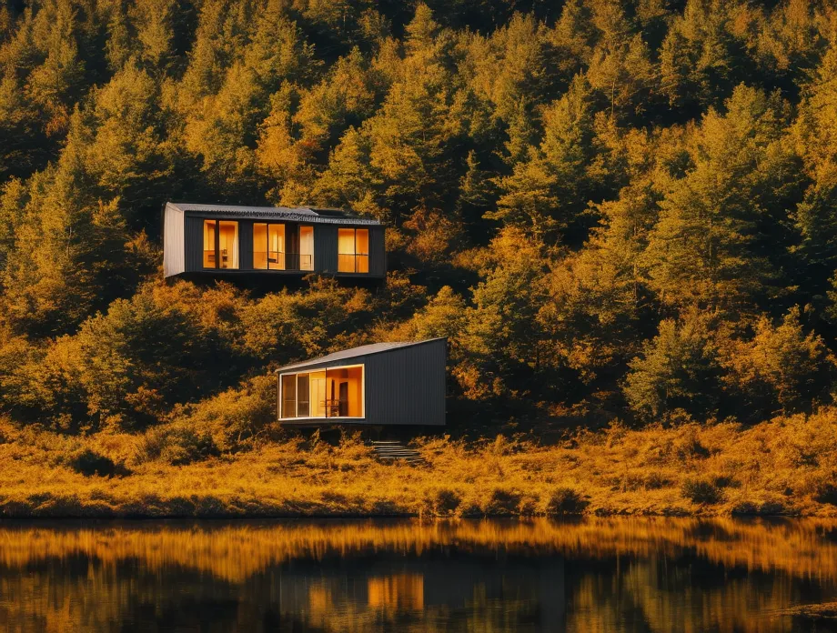 La imagen muestra dos cabañas modernas en el bosque. Las cabañas son de color negro y tienen grandes ventanas. Están rodeadas de árboles altos y las hojas se están volviendo amarillas y naranjas. Hay un lago frente a las cabañas y el agua está reflejando los colores de los árboles. El cielo es azul y hay algunas nubes. Las cabañas están cerca una de la otra.