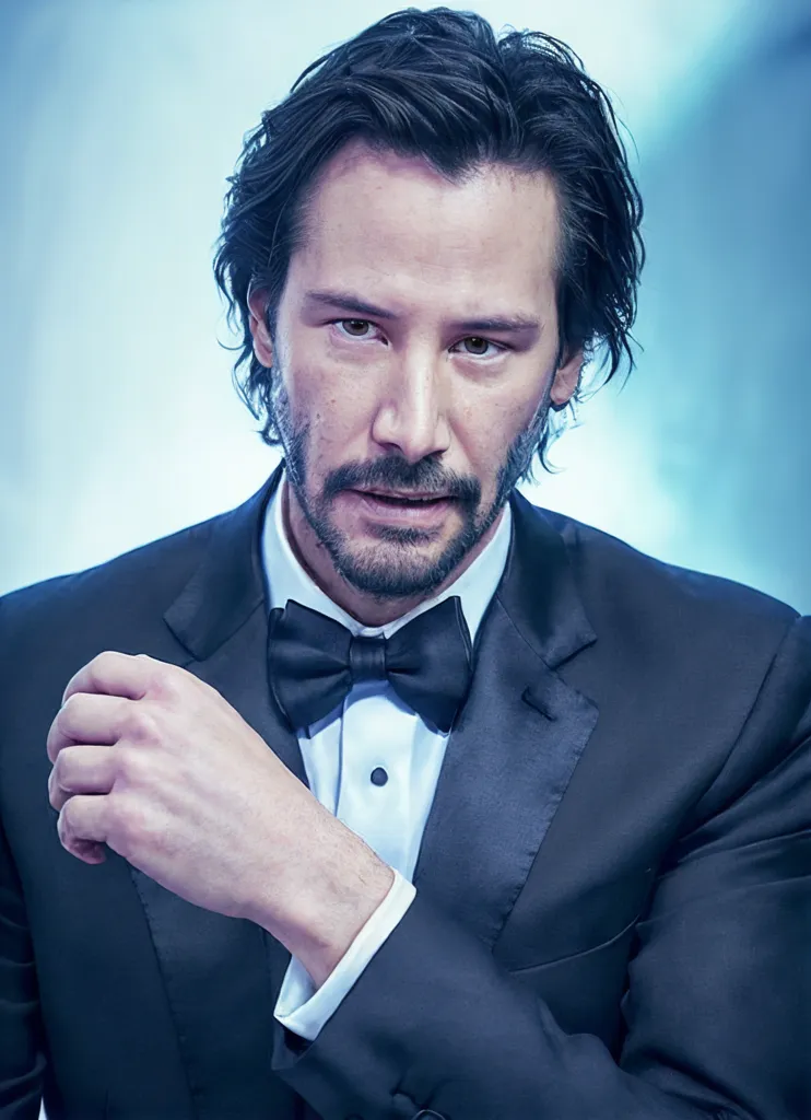 The image shows Keanu Reeves, a Canadian actor. He is wearing a black suit and a bow tie. His hair is long and dark, and he has a beard. He is looking at the camera with a serious expression.