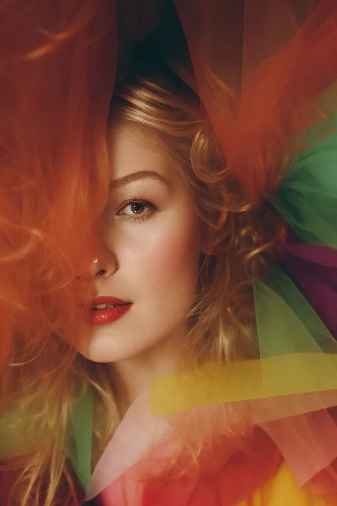 The image is a portrait of a young woman with blonde hair and red lipstick. She is wearing a colorful dress with a plunging neckline. The woman is standing in front of a white background, and her face is partially obscured by a sheer red scarf. The scarf is draped over her head and shoulders, and it creates a sense of mystery and intrigue. The woman's eyes are looking directly at the viewer, and she has a slightly defiant expression on her face. The image is taken from a close-up angle, and it captures the woman's beauty and vulnerability.