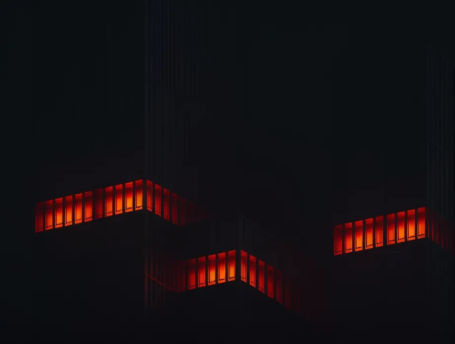 L'image représente un bâtiment sombre avec des fenêtres rouges illuminées la nuit. Le bâtiment est grand et a beaucoup de fenêtres. Les fenêtres sont disposées de manière régulière. Le bâtiment est au centre de l'image et est entouré par l'obscurité. L'image est très sombre et a une atmosphère mystérieuse.