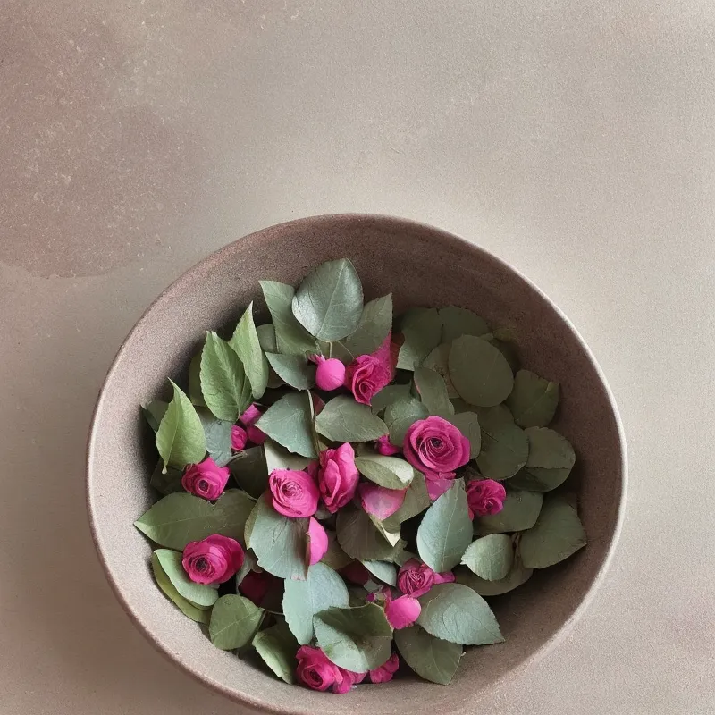 The image is a close-up of a bowl filled with pink roses and green leaves. The bowl is placed on a solid surface. The roses are small and delicate, with a light pink color. The leaves are small and oval-shaped, with a deep green color. The bowl is rustic and has a rough texture. The background is a solid color, which makes the bowl and its contents stand out.