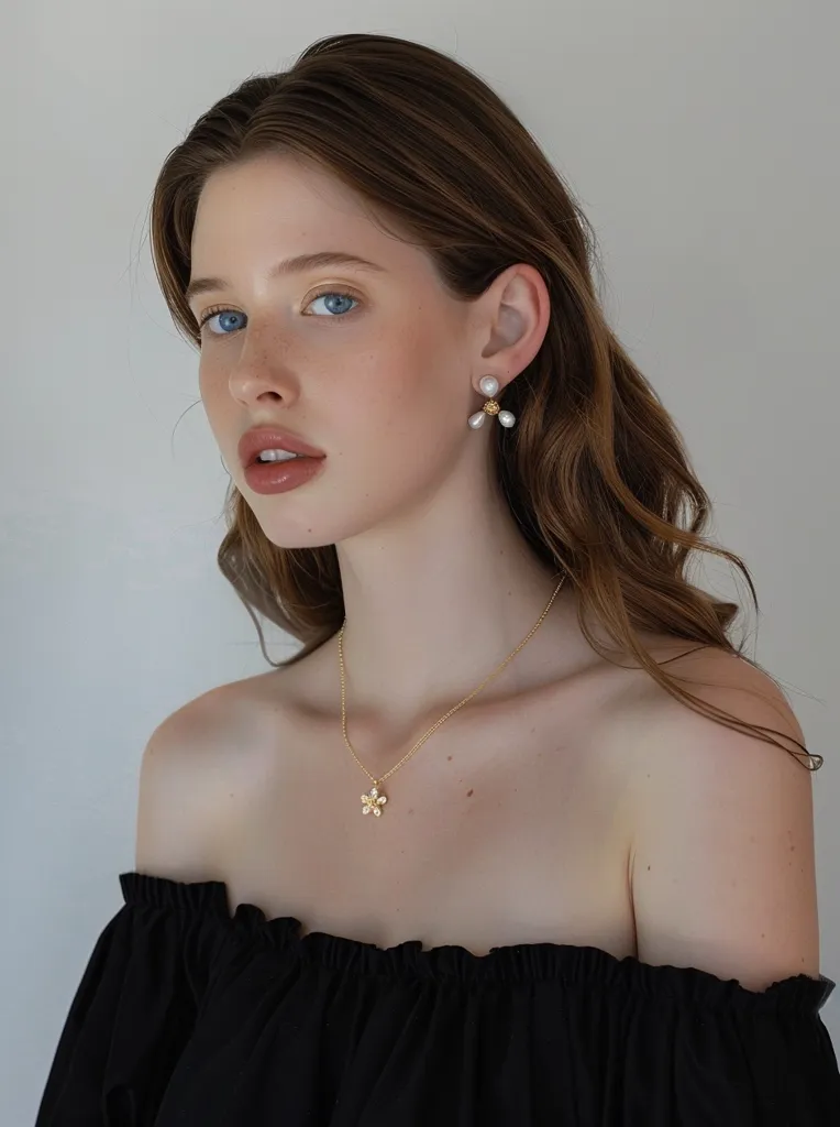 The image shows a young woman with long brown hair and blue eyes. She is wearing a black off-the-shoulder top and a gold necklace with a flower-shaped pendant. She is also wearing gold earrings with three pearls each. The woman has a soft smile on her face and is looking at the camera. She is standing in front of a white background.