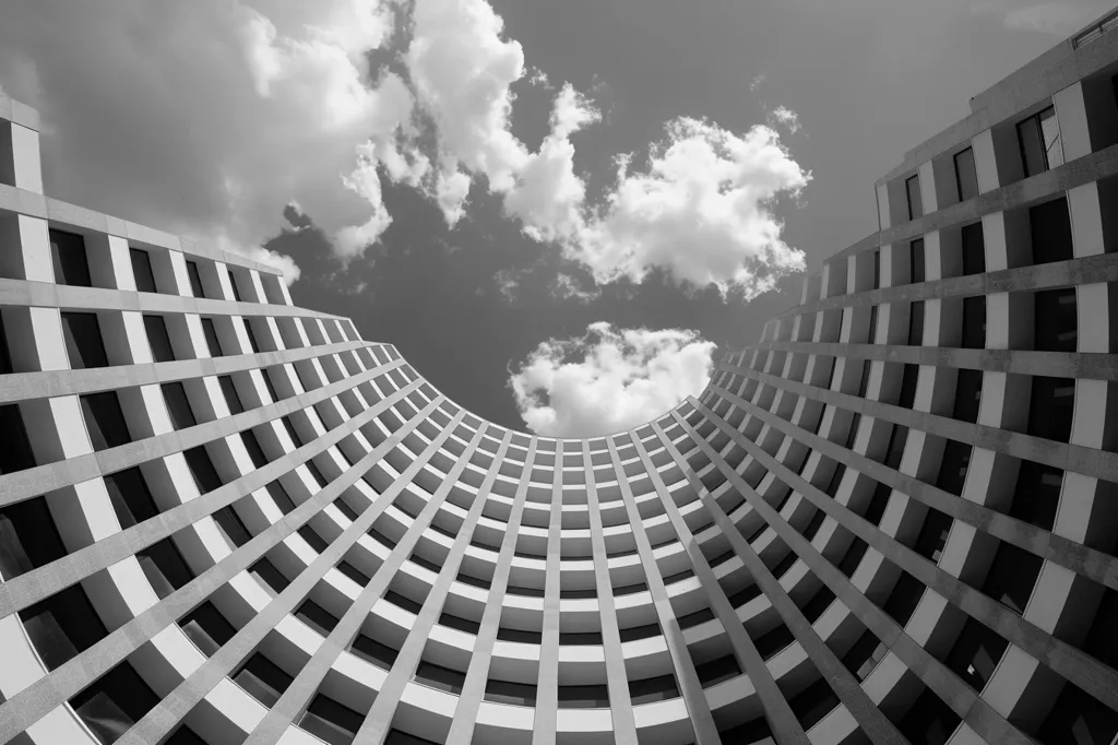 A black and white photo of a modern building with a curved facade, looking up from the ground. The building is made up of a grid of windows and concrete, creating a strong geometric pattern. Above the building, fluffy clouds are visible against a bright sky. The photo captures the interplay of man-made structures and natural elements.