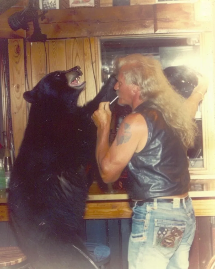 A man with long blonde hair and a leather vest is standing in a bar with a large black bear. The man is holding a stick in his hand and is looking at the bear. The bear is looking at the man and has its mouth slightly open. The bar is made of wood and has a wooden counter. The man and the bear are both in the center of the image.