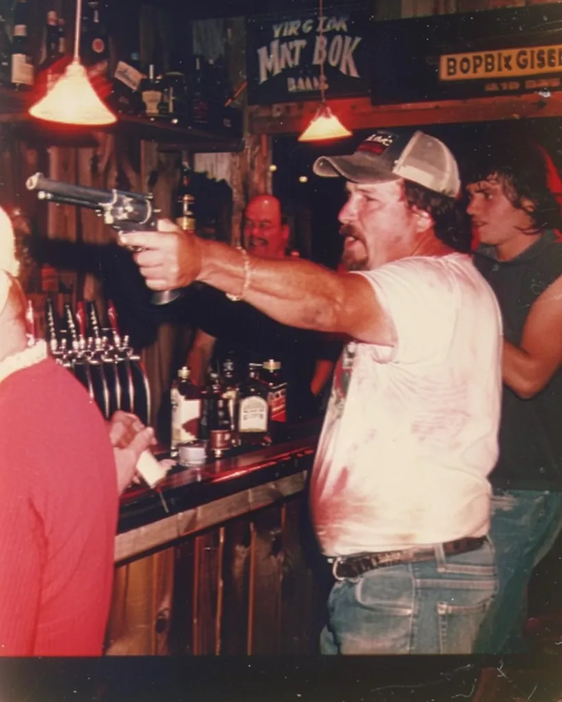 A man in a white t-shirt and a baseball cap is holding a gun and pointing it at the camera. He is standing at a bar with other men behind him. The man is looking at the camera with a serious expression on his face. There are bottles of alcohol on the bar counter and a sign for a bar behind him. The photo is a bit blurry and grainy.