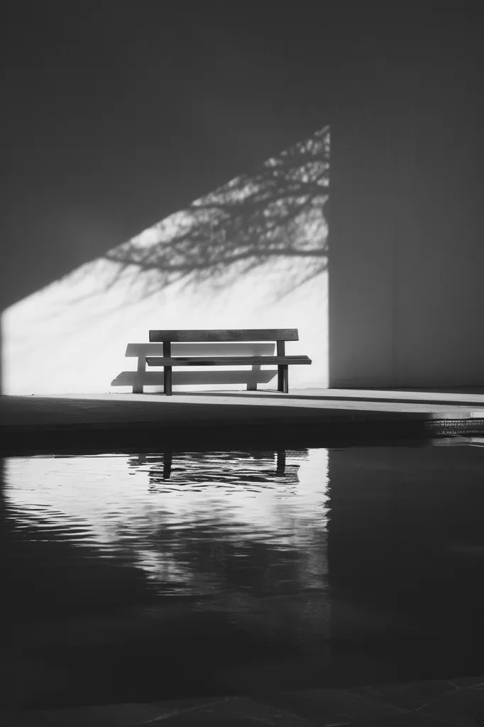 A black and white photograph of a lone bench sitting in front of a body of water.  The bench is made of metal and has two seats.  The bench is casting a shadow on the ground in front of it.  The water is reflecting the light from the sky, creating a beautiful, tranquil scene.  The image is devoid of people, creating a sense of solitude and peace.