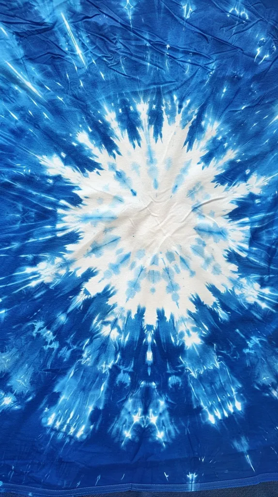 The image shows a close-up of a blue and white tie-dye pattern. The fabric has a circular design, with a white center surrounded by a ring of blue, then a ring of white, and finally another ring of blue. The pattern creates a swirling effect, like a vortex of color. The fabric appears to be slightly wrinkled, adding to the organic, hand-crafted feel of the tie-dye.