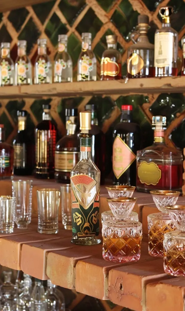 A wooden shelf displays a variety of liquor bottles,  with intricate labels and  gold accents. Some bottles have a clear glass, others are brown or green.  In the foreground, several clear glass tumblers and glasses with a  gold rim are arranged on  a brick-like surface. The scene evokes a sense of classic elegance and timeless sophistication,  perfect for a luxurious bar or home setting.
