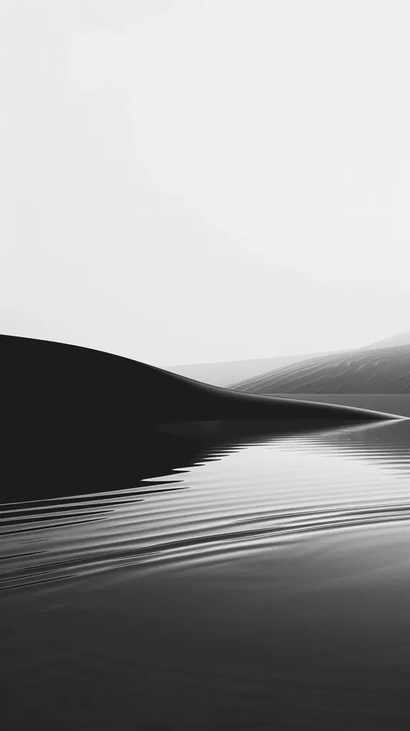 A black and white photograph of a calm body of water with a gentle ripple pattern. In the distance, a dark hill is visible against a hazy sky. The image captures a serene and minimalist landscape, with soft lines and muted tones. The water's surface reflects the sky, creating a sense of tranquility and vastness.