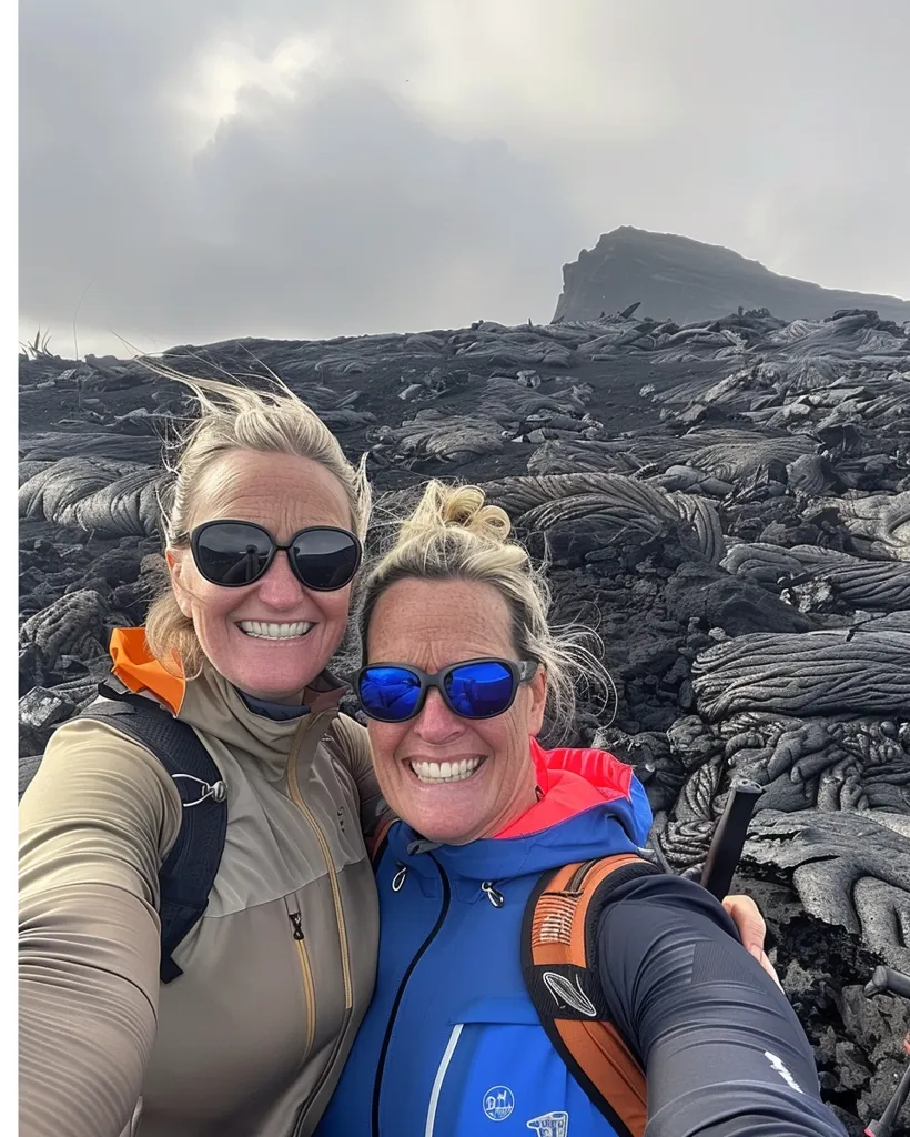 Two women wearing sunglasses and hiking gear smile for a selfie.  They stand in front of a field of cooled lava rock.  Behind them is a volcanic peak in the distance.  The sky is cloudy, giving the scene a dramatic, yet beautiful feel.