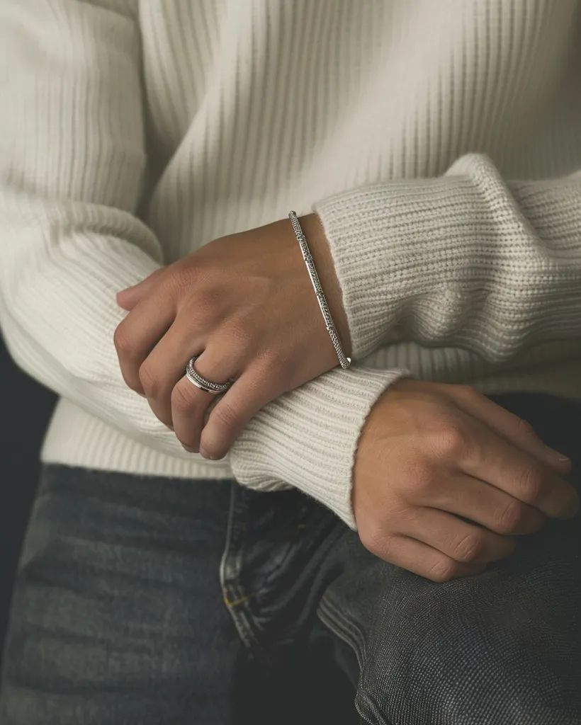 A person wearing a white sweater and blue jeans is sitting with their arms crossed. The left hand is resting on the right arm, and is wearing a silver bracelet and ring.  The image is cropped at the elbows and above the knees.  The background is out of focus.