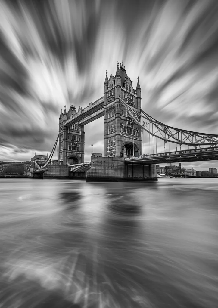 A black and white photograph of Tower Bridge in London, England. The bridge is captured in sharp detail, while the sky and water are blurred, creating a sense of motion. The iconic towers and suspension cables are visible, and the image is taken from a low angle, emphasizing the bridge's grandeur. The cloudy sky adds a dramatic touch to the composition.
