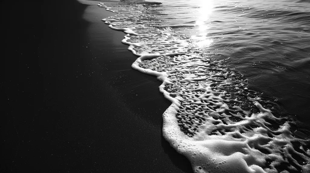 A black and white image captures the contrast between a dark sandy beach and the foamy white waves rolling in from the ocean. The waves are textured and detailed, with the light reflecting off the water, creating a beautiful scene. The image is shot from a low angle, emphasizing the scale of the waves and the endless expanse of the ocean.