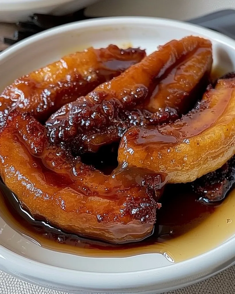 A white bowl holds a generous serving of caramelized bananas. The bananas are cut into thick slices, coated in a rich, dark brown sauce, and glistening with a sweet glaze. They appear soft and tender, with a slightly crisp exterior. The sauce pools at the bottom of the bowl, offering a visually appealing contrast to the golden brown bananas.