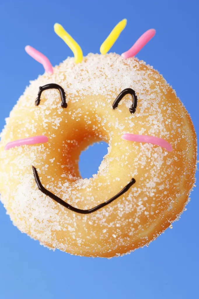 A sugary donut with a smiling face drawn on it. It has pink and yellow icing and is covered in sugar. The donut is against a blue background.