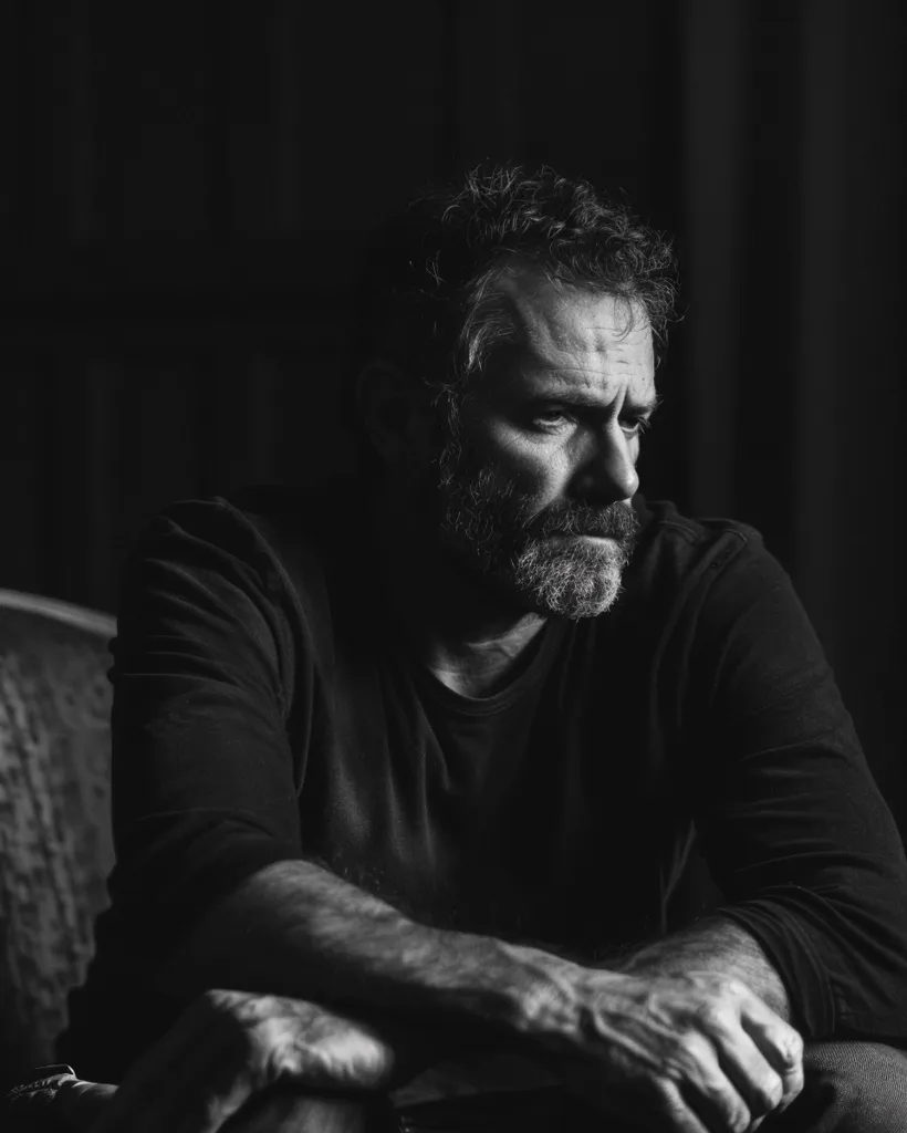 A man with a beard and a pensive expression sits in a chair. The image is in black and white, creating a moody and atmospheric feel. He is wearing a dark shirt and his arms are crossed, suggesting a sense of contemplation or introspection. The shadows and low-key lighting create a sense of depth and mystery, adding to the overall mood of the image.  The subject's gaze is directed downward, further emphasizing his thoughtfulness.