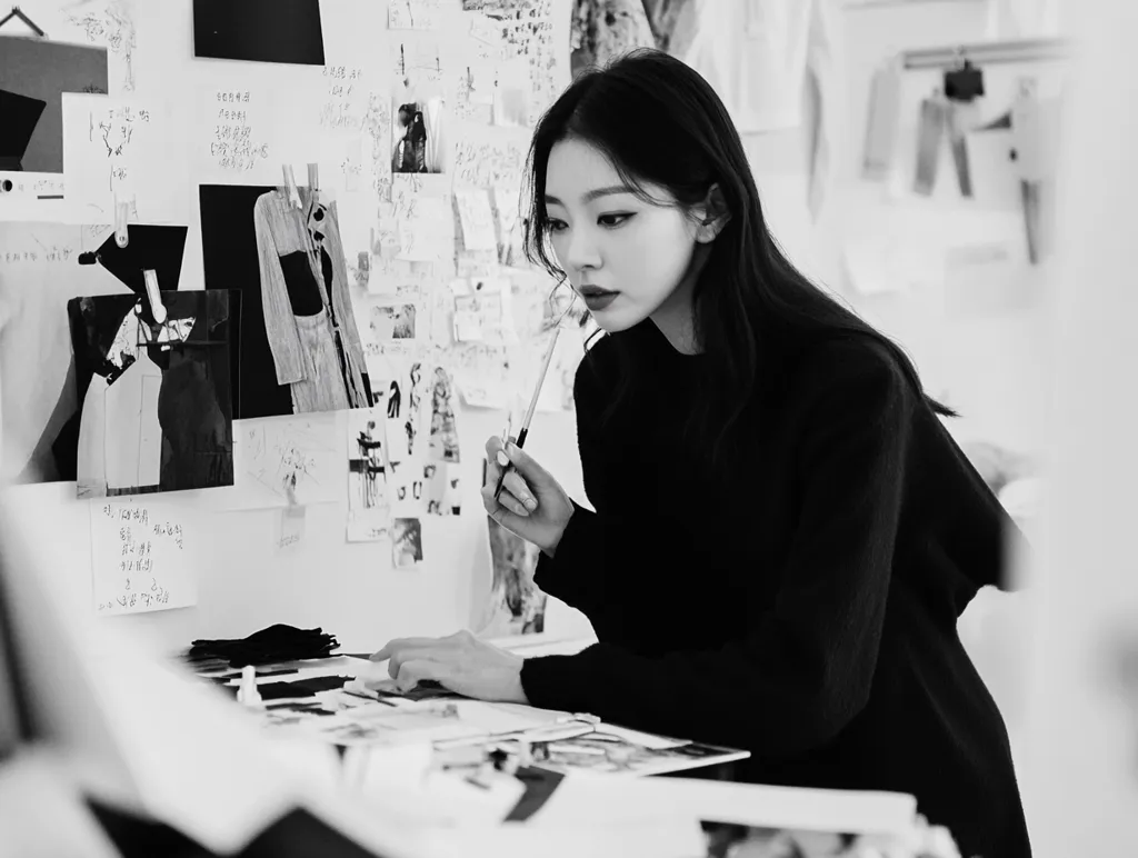 A woman with long dark hair, wearing a black sweater, sits at a table covered in papers and sketches. She is holding a pencil and looking down at her work. The wall behind her is covered in notes, sketches, and fabric swatches. The photo is in black and white, creating a moody and artistic feel.