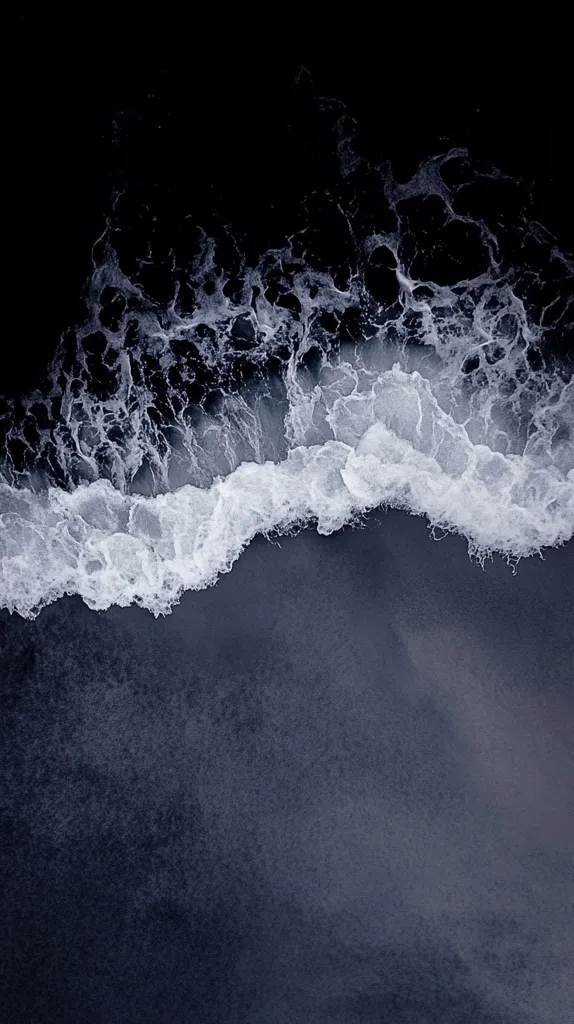 An aerial shot of a dark beach with a white wave crashing onto the shore. The wave's foam creates a stark contrast against the dark sand, leaving an impression of the wave's power and movement. The image is captured with a high-contrast, dramatic effect.