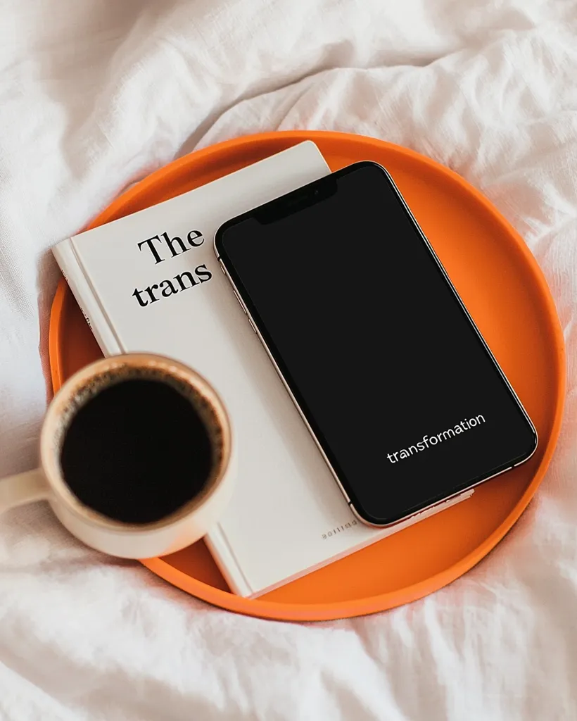A white book with the words "The transformation" on it lies in an orange tray. A smartphone with a black screen sits on top of the book. A white cup with coffee rests on the tray. All are on a white bedsheet with wrinkles. The image suggests a moment of calm and reflection, perhaps before a journey of self-improvement.