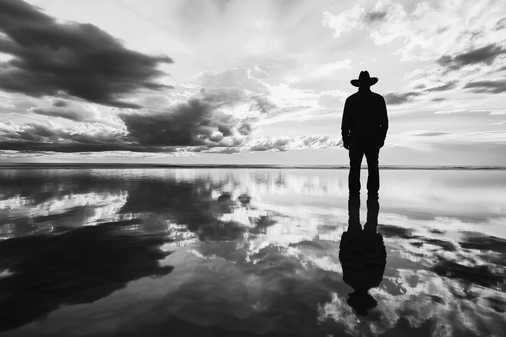 A solitary figure, silhouetted against a cloudy sky, stands on a vast, still body of water.  Their cowboy hat casts a shadow as they gaze out at the horizon. The water reflects the clouds above, creating a mirrored image of the scene. The image is a study in contrasts, with the darkness of the figure and clouds against the lightness of the water and sky.  It evokes a sense of isolation and contemplation, as if the figure is lost in thought.