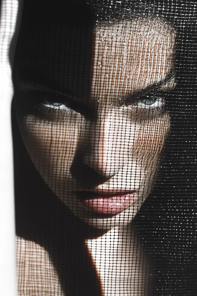 A woman's face is obscured by a black net. Her piercing blue eyes are visible through the mesh. Her lips are painted a vibrant red. The lighting is dramatic, creating deep shadows and highlights. The image is stark and mysterious, suggesting a sense of intrigue and allure.