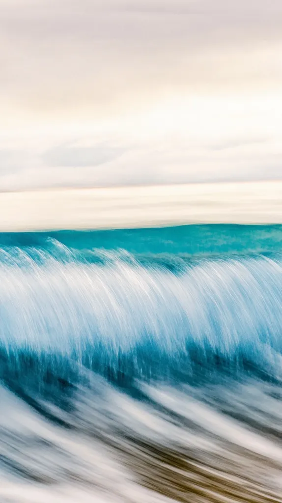 The image captures a wave crashing on the shore, with a focus on the motion blur effect created by the movement of the water. The white, foamy crest of the wave contrasts with the deep blue of the water below. The background features a soft, white sky, suggesting a peaceful and tranquil setting. The overall image evokes a sense of dynamism and the power of nature.