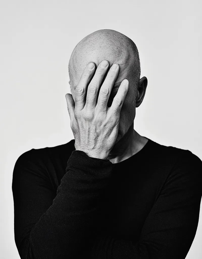 A black and white photograph of a man with his hand covering his face. He is wearing a black long-sleeved shirt. His face is obscured, leaving the viewer to imagine his emotions. The image conveys a sense of sadness, frustration, or perhaps even shame. The stark contrast between the light and dark tones emphasizes the subject's solitary nature. The photograph suggests a feeling of introspection and a desire to hide from the world.