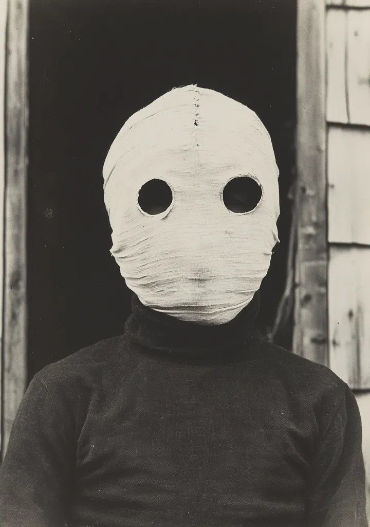 A black and white photo of a person wearing a white cloth mask covering their face, leaving only their eyes visible. The mask is tightly wrapped around the head, and the person is wearing a dark turtleneck sweater. The background is blurred, but a wooden door or wall is visible. The image has a haunting and mysterious feel.