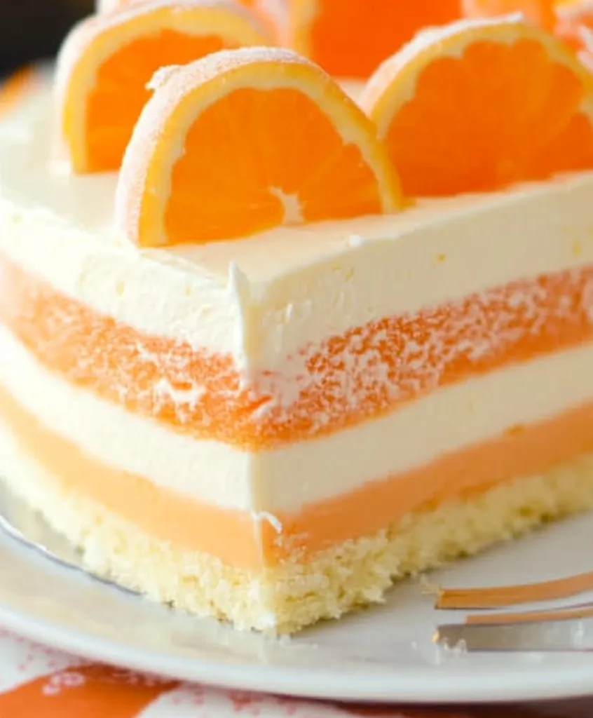 A slice of layered cake is decorated with orange slices. The cake has alternating layers of white and orange, and the bottom layer is a crumb crust. The cake is on a white plate and a fork is beside it. The image is close-up and captures the texture of the cake.