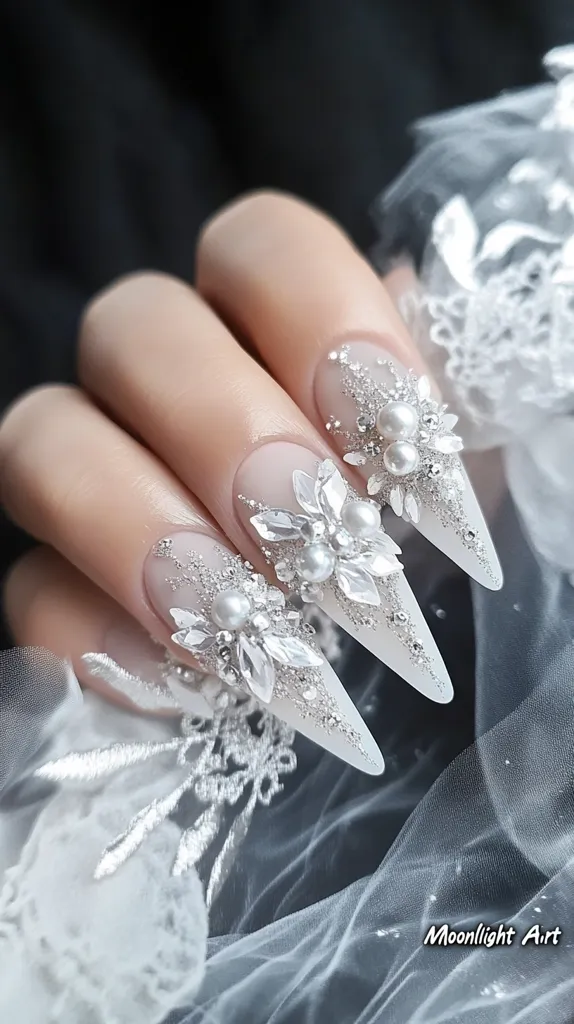 A close-up of a hand with long, pointed nails decorated with intricate designs made of silver glitter, rhinestones, and pearls. The nails are painted a light, almost translucent white, and the hand is resting on a sheer white fabric, creating a delicate and elegant image. The image evokes a sense of femininity, sophistication, and bridal glamour.