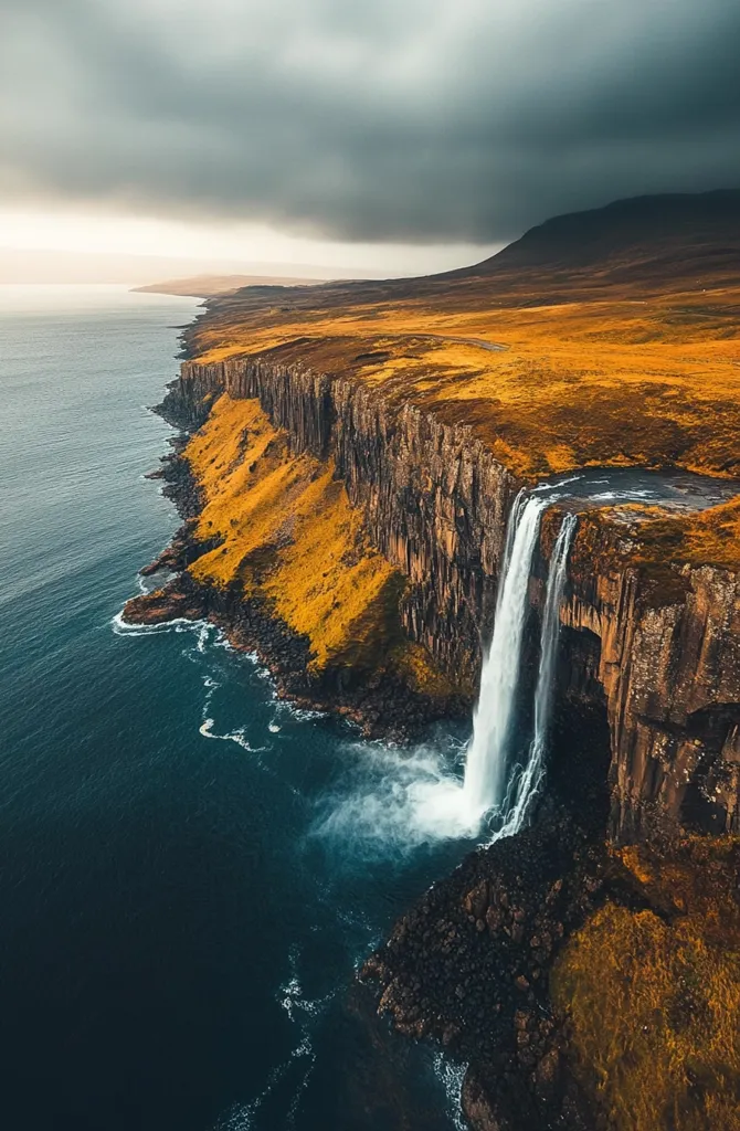 A dramatic aerial view of a waterfall cascading down a sheer cliff face into the ocean. The cliff is covered in a mix of green vegetation and dark rock, and the ocean is a deep blue. A storm cloud hangs in the sky, casting a shadow over the landscape. The scene is both majestic and powerful.