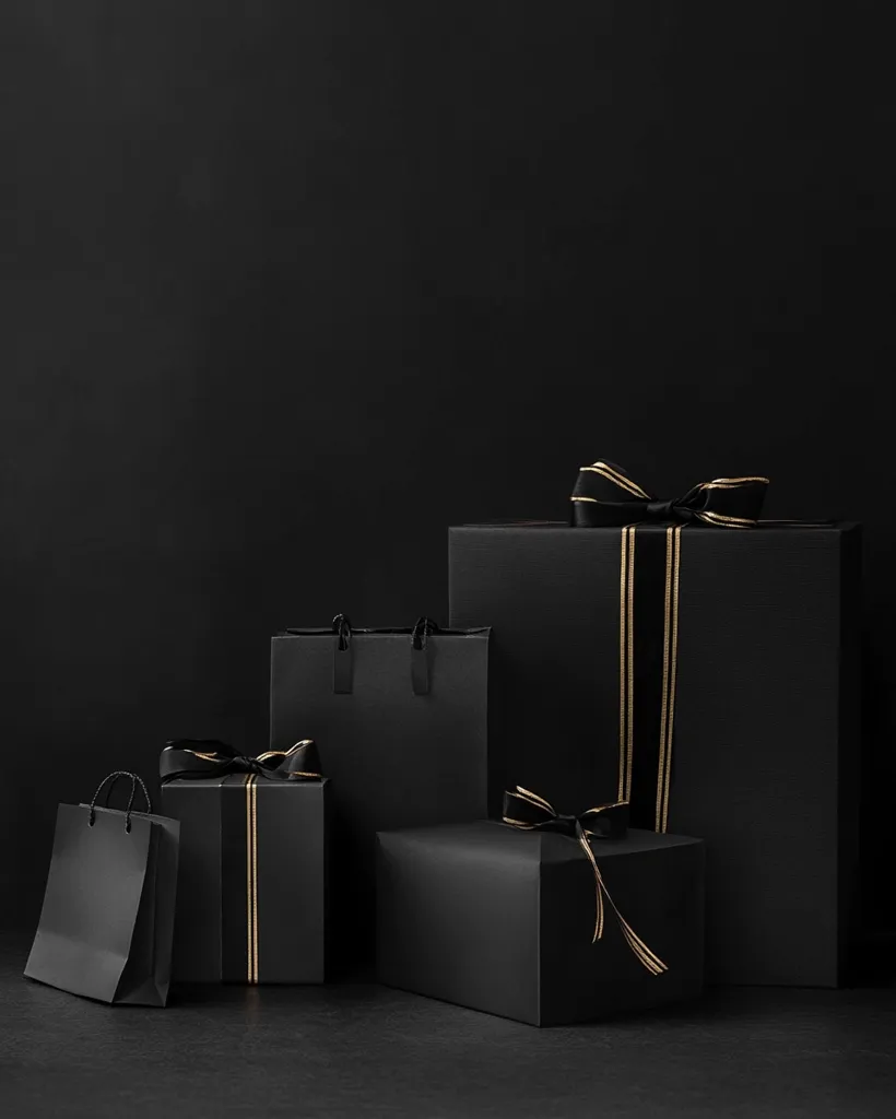 Four black gift boxes sit on a black surface, all tied with black ribbon and gold trim. One box is a shopping bag, another is tall and narrow, and the two largest boxes have a large bow on top. They are all set against a black backdrop. The image creates a stark and minimalist aesthetic, with the black and gold highlighting a sense of luxury and elegance.