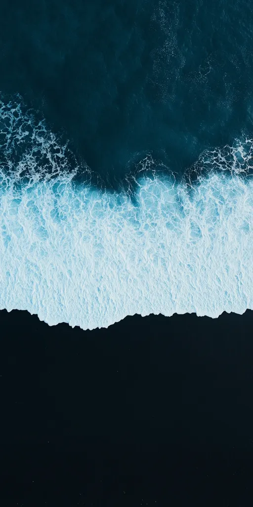 An aerial view of a black sand beach meeting the ocean. White foamy waves crash against the shore creating a striking contrast with the dark sand and the deep blue ocean.  The image captures a moment of serenity and natural beauty.
