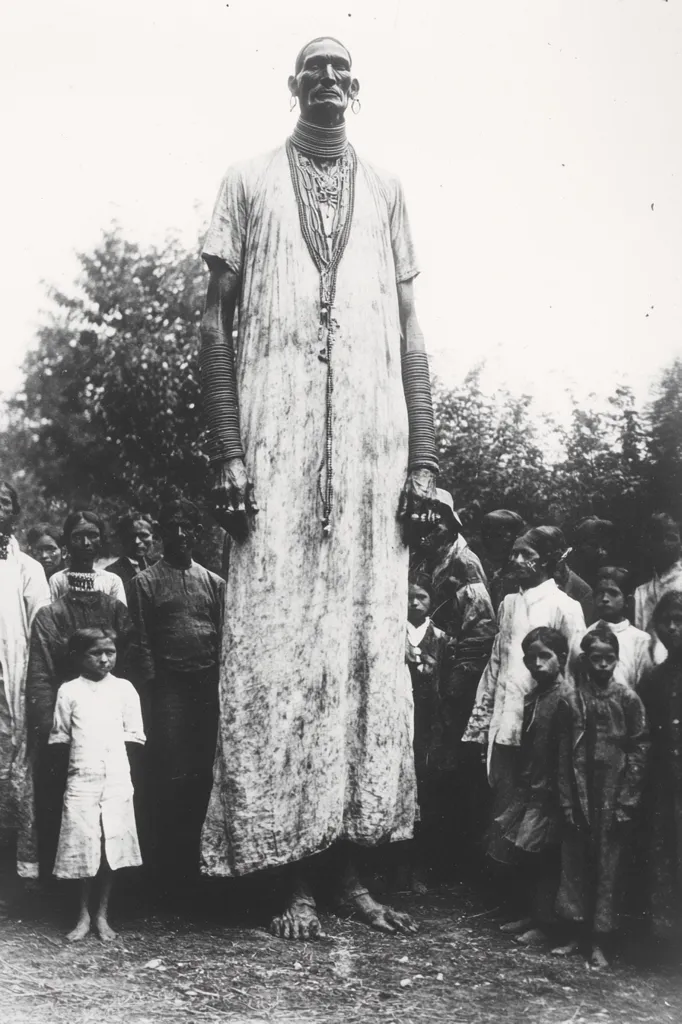 A tall woman, adorned with necklaces and bracelets, stands amidst a group of children and adults. She wears a long, flowing gown that obscures her feet, emphasizing her imposing height. The photo captures a sense of curiosity and awe, as the children gaze up at the woman with wonder. The black and white tones lend a timeless quality to the image, suggesting a bygone era.