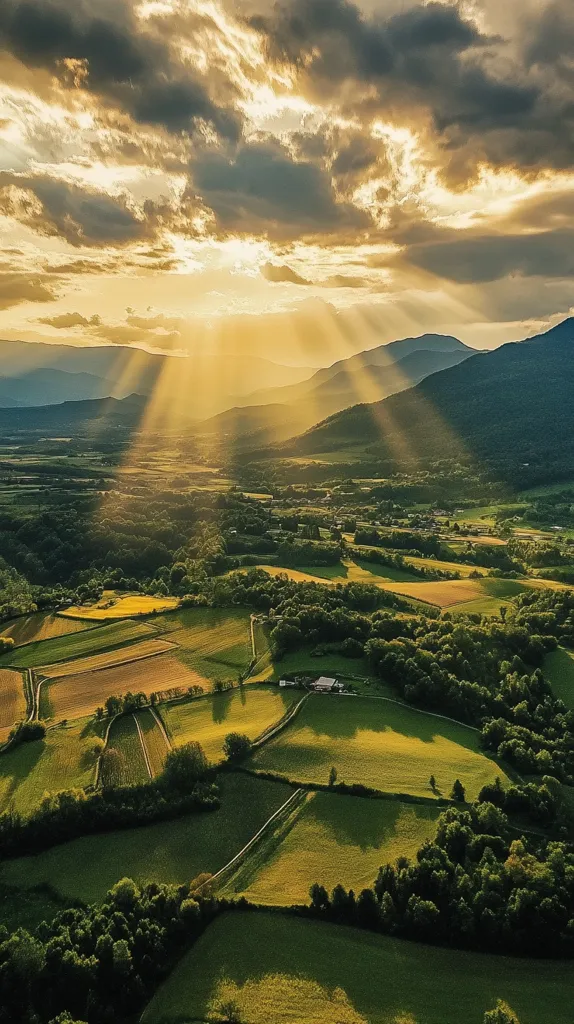 The image captures a breathtaking aerial view of a rolling countryside bathed in the golden light of a setting sun.  Rays of sunlight pierce through the clouds, casting long shadows over the lush green fields and forests below.  The distant mountains rise majestically in the background, creating a picturesque and serene landscape.  The scene evokes a sense of peace and tranquility, with nature's beauty on full display.