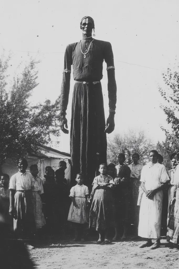 A very tall man stands in the center of the image, towering over a group of children and adults. He is wearing a long robe and a necklace, and has a serious expression on his face. The children are dressed in simple clothing and look up at the man in awe. The adults are also looking at the man, but their expressions are more reserved. The scene is set in an outdoor location, with trees in the background. The image is in black and white, and the lighting is soft.  The overall tone of the image is one of wonder and respect.