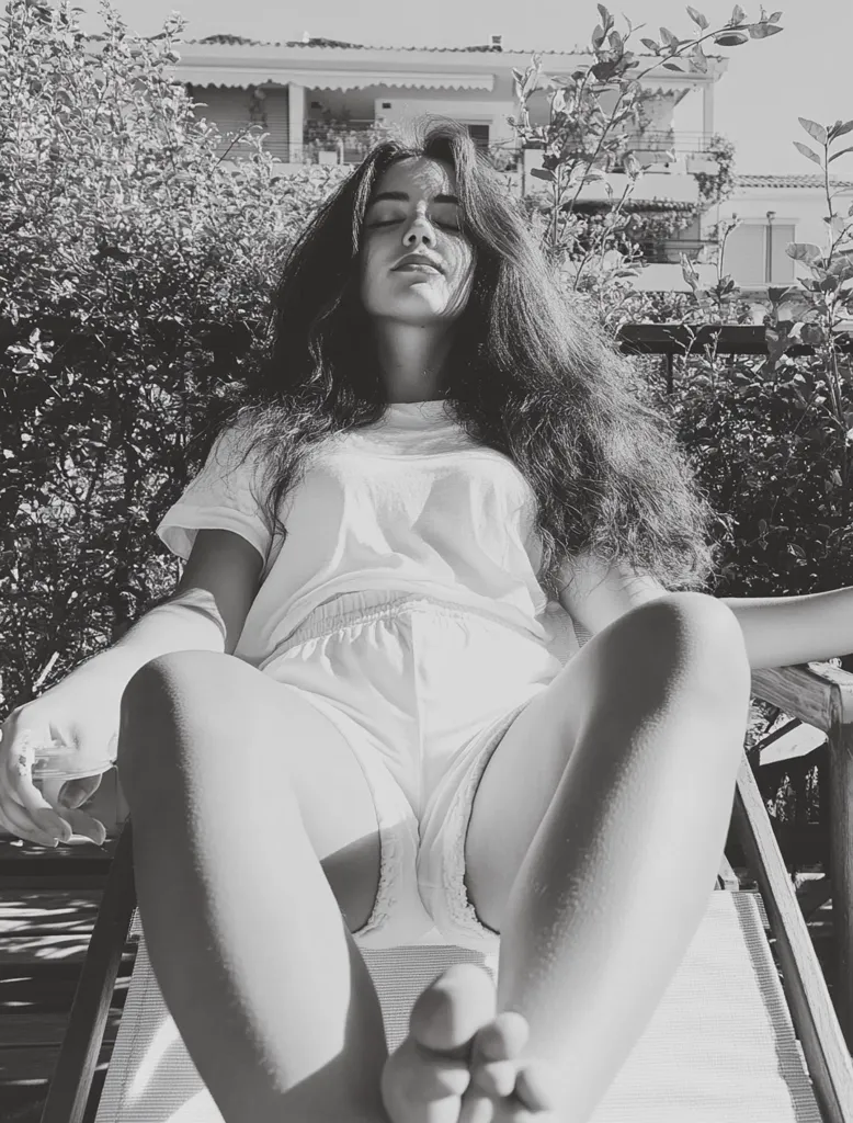 A young woman with long, dark hair is sitting on a chair, her eyes closed and her head tilted back. She is wearing a white t-shirt and shorts, and her legs are stretched out in front of her. The photo is in black and white, and the woman's relaxed posture suggests a sense of peace and tranquility. The lush greenery behind her adds to the peaceful atmosphere.  The photo evokes a sense of serenity and calm.