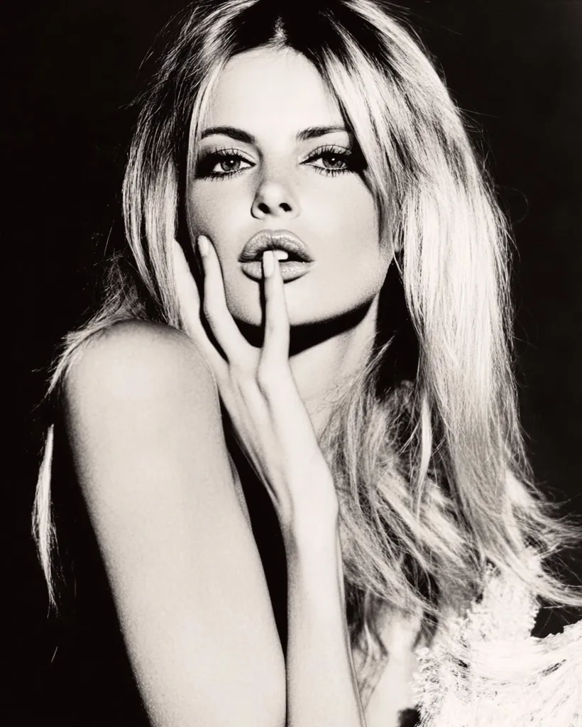 A black and white portrait of a woman with long, flowing hair. She is looking directly at the camera with a slightly parted mouth and her finger pressed against her lips. She has a soft, ethereal look, with her hand resting on her cheek. The image is soft-focus and has a romantic and timeless feel.