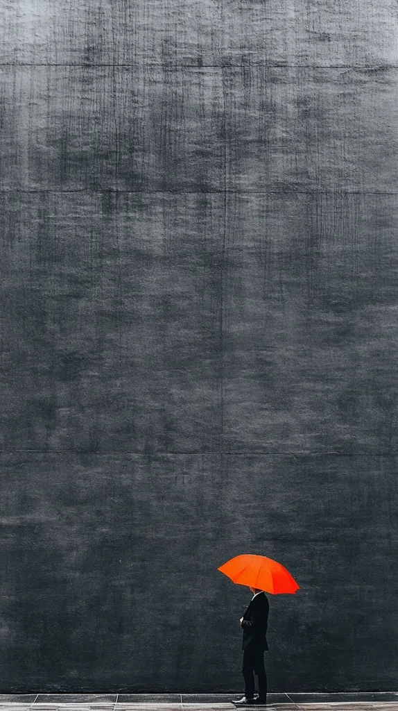 A man in a black suit stands holding a bright red umbrella in front of a plain, grey wall. The man is facing away from the camera, with his head slightly tilted downwards. The image is taken from a low angle, creating a sense of perspective and emphasizing the man's small stature against the vast wall. The scene is simple and minimalist, focusing on the contrast between the bright color of the umbrella and the subdued background.