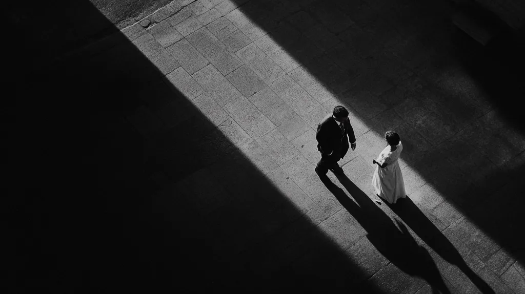 A black and white photograph taken from a high angle shows a man and woman walking in opposite directions on a paved surface. The man wears a suit and the woman is in a white dress. The sun casts a long shadow from a building onto the ground.  The image is stark and minimalist, with strong contrasts between light and shadow.  The figures appear small and distant, creating a sense of loneliness and isolation.