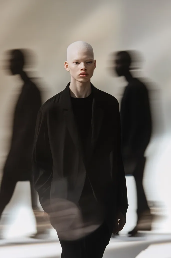 A bald man wearing a black blazer and dark pants stands in the foreground. The background is blurry with two indistinct figures. The image has a stark, minimalist aesthetic. The subject's expression is neutral, creating a sense of mystery. The lighting is soft and diffused. The overall composition is simple and elegant.