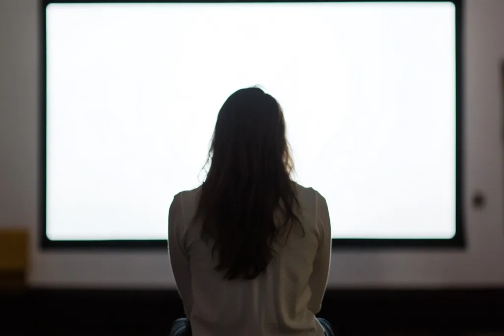 A woman sits in front of a large, blank screen, her silhouette outlined against the bright white light. The screen is a television or projector, and the woman is likely watching a movie or presentation. The image creates a sense of anticipation and mystery.