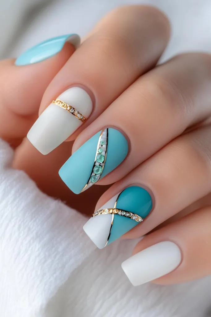 A hand with manicured nails painted in a blue and white color scheme with gold accents. The nails are decorated with geometric patterns and rhinestones. The hand is resting on a white surface. The image focuses on the intricate details of the nail art.