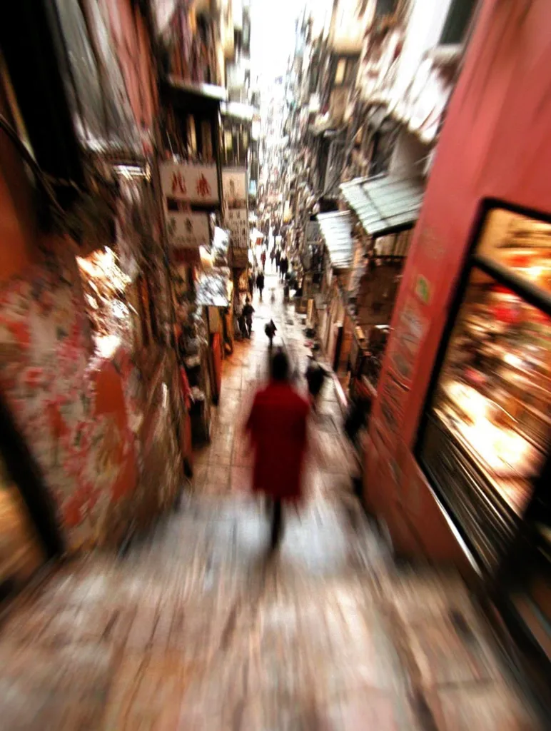 The photo captures a narrow, bustling street in Hong Kong, a blur of motion and light. Buildings on either side press in, their facades painted in warm tones. A solitary figure in red walks through the crowd, their form indistinct amidst the blur. The scene is dynamic, conveying the energy and intensity of urban life.