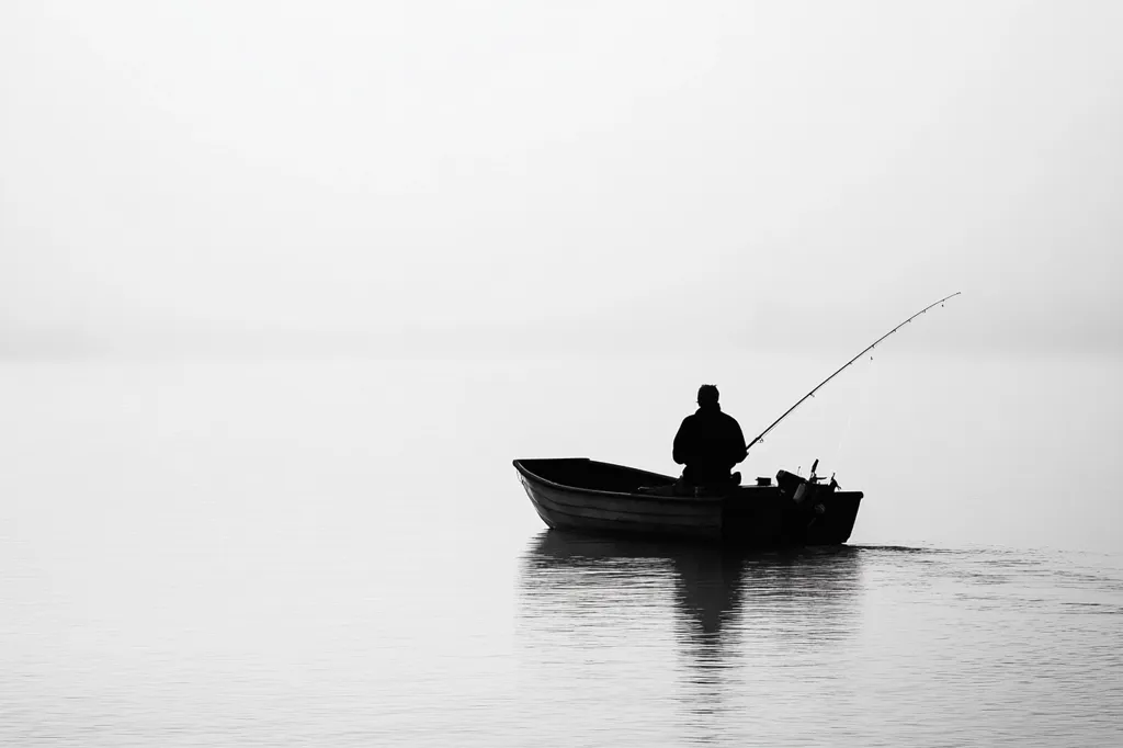 A lone fisherman sits in a rowboat, casting his line into the calm, still water.  The fog is thick and the sky is overcast, creating a moody, atmospheric scene.  The image is black and white, emphasizing the stark contrast between the figure and the empty surroundings.  The silence and solitude of the moment are palpable.