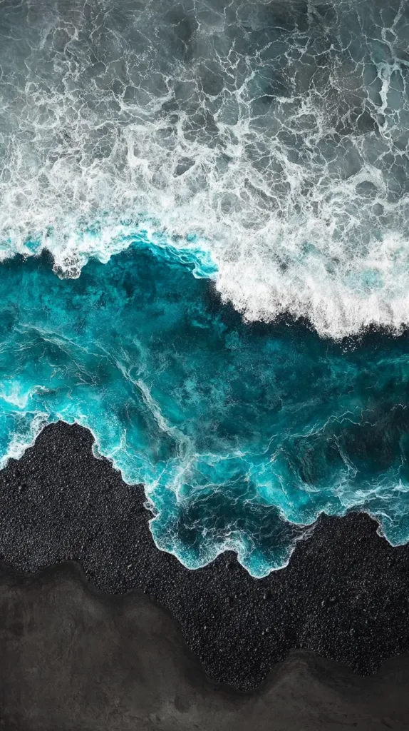 An aerial view of a black sand beach with turquoise ocean waves crashing on shore. The water is a vibrant blue, and the waves are capped with white foam. The black sand creates a stark contrast against the blue water, creating a visually striking scene. The image captures the raw power and beauty of nature.