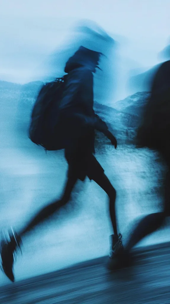 A blurred silhouette of a person wearing a backpack is running through a mountainous landscape. The image is shot in a blue hue, creating a sense of mystery and motion. The person's figure fades into the background, leaving the viewer with a sense of intrigue. The image could be interpreted as a representation of travel, adventure, or even a fleeting moment in time.